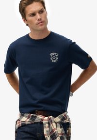 Hombre que lleva una camiseta azul marino de Superdry, una camisa de cuadros atada alrededor de la cintura, jeans oscuros y un reloj en la muñeca, mirando hacia la izquierda.