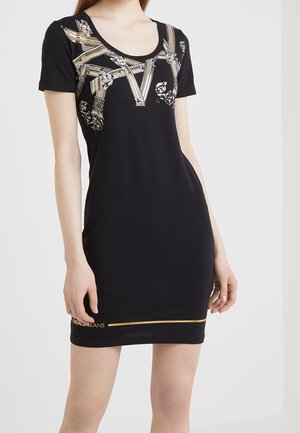 Femme portant une courte robe noire avec un imprimé baroque doré sur le haut et une ligne dorée avec un texte près de l'ourlet, debout devant un fond uni.