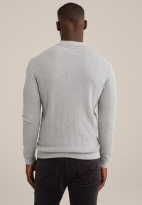 Muž nosící světle šedý žebrovaný svetr s texturou a tmavé kalhoty, zobrazený zezadu na neutrálním jednobarevném pozadí.