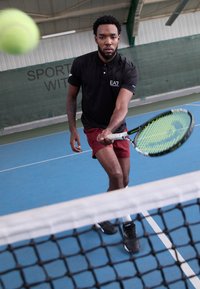 Tennisspieler in einem schwarzen Polohemd und kastanienbraunen Shorts, der einen grünen Tennisball mit einem Schläger auf einem blauen Indoorplatz schlägt.