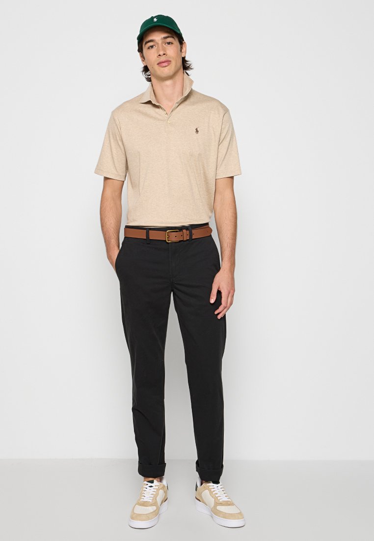 Polo beige avec un petit logo, pantalon noir, ceinture marron et baskets beiges avec des accents blancs. Design simple, tenue décontractée.