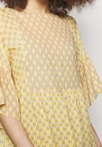 Beżowa i żółta sukienka w wzory o luźnym kroju, z krótkimi rozkloszowanymi rękawami oraz teksturowanym geometrycznym wzorem. Miękka, lekka tkanina.