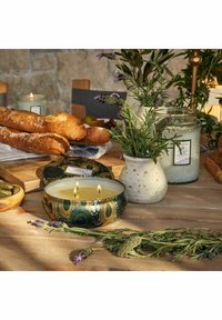 Doftljus i en rund, mönstrad behållare med två veker; omgiven av örter, en texturerad vas och brödlimpor på ett träbord.