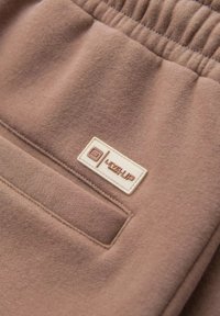 Pantaloni della tuta color marrone chiaro con una vita elastica, morbida al tatto, e un'etichetta rettangolare bianca con il marchio "LEVEL UP" sulla tasca.