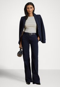 Žena v námorníckom modrom blejzri prehodenom cez ramená, bielom a námorníckom pruhovanom topu, námorníckych širokých nohaviciach, držiaca malú čiernu kabelku.