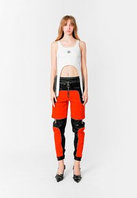 Joven mujer vestida con un top blanco sin mangas y recortado con una abertura y pantalones de cremallera rojos y negros, de pie frente a un fondo blanco liso.
