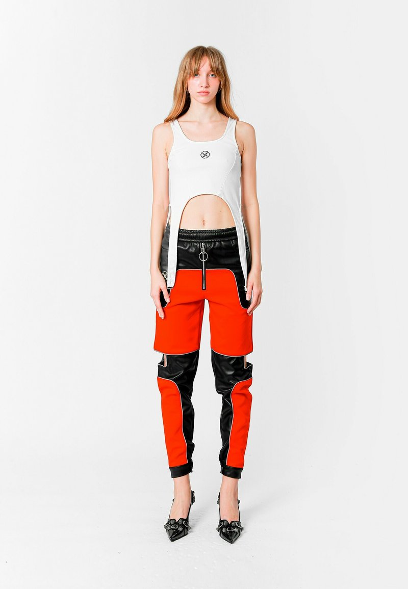 Joven mujer vestida con un top blanco sin mangas y recortado con una abertura y pantalones de cremallera rojos y negros, de pie frente a un fondo blanco liso.