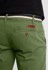 Pantalon en coton vert avec une texture lisse, présentant une étiquette de marque en cuir marron et une ceinture tissée multicolore. Détail de poche arrière visible.