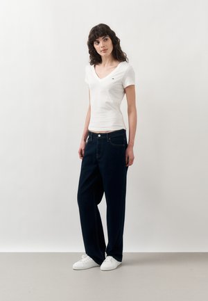 Γυναίκα με σκούρα σγουρά μαλλιά, που φοράει λευκό T-shirt με V-λαιμόκοψη, άνετο σκούρο τζιν και λευκά αθλητικά, στέκεται μπροστά σε απλό λευκό φόντο.