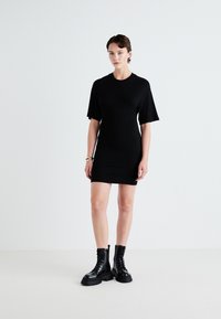 Robe noire moulante avec des manches courtes et un col rond. Portée avec des bottes de combat noires à lacets, mettant en valeur une silhouette élégante et ajustée.