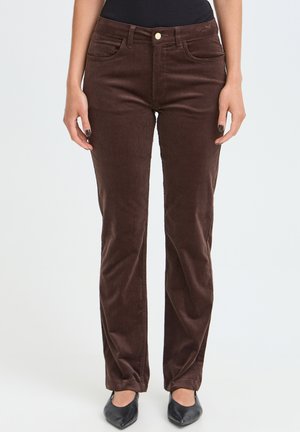 Femme portant un pantalon en velours côtelé marron foncé à jambes droites et des chaussures noires à bouts pointus, debout devant un fond clair uni.