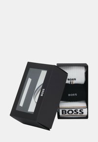 Schwarze Box mit transparentem Deckel, die drei Paar weiße und schwarze Socken mit der Marke "BOSS" enthält, gestapelt darin.