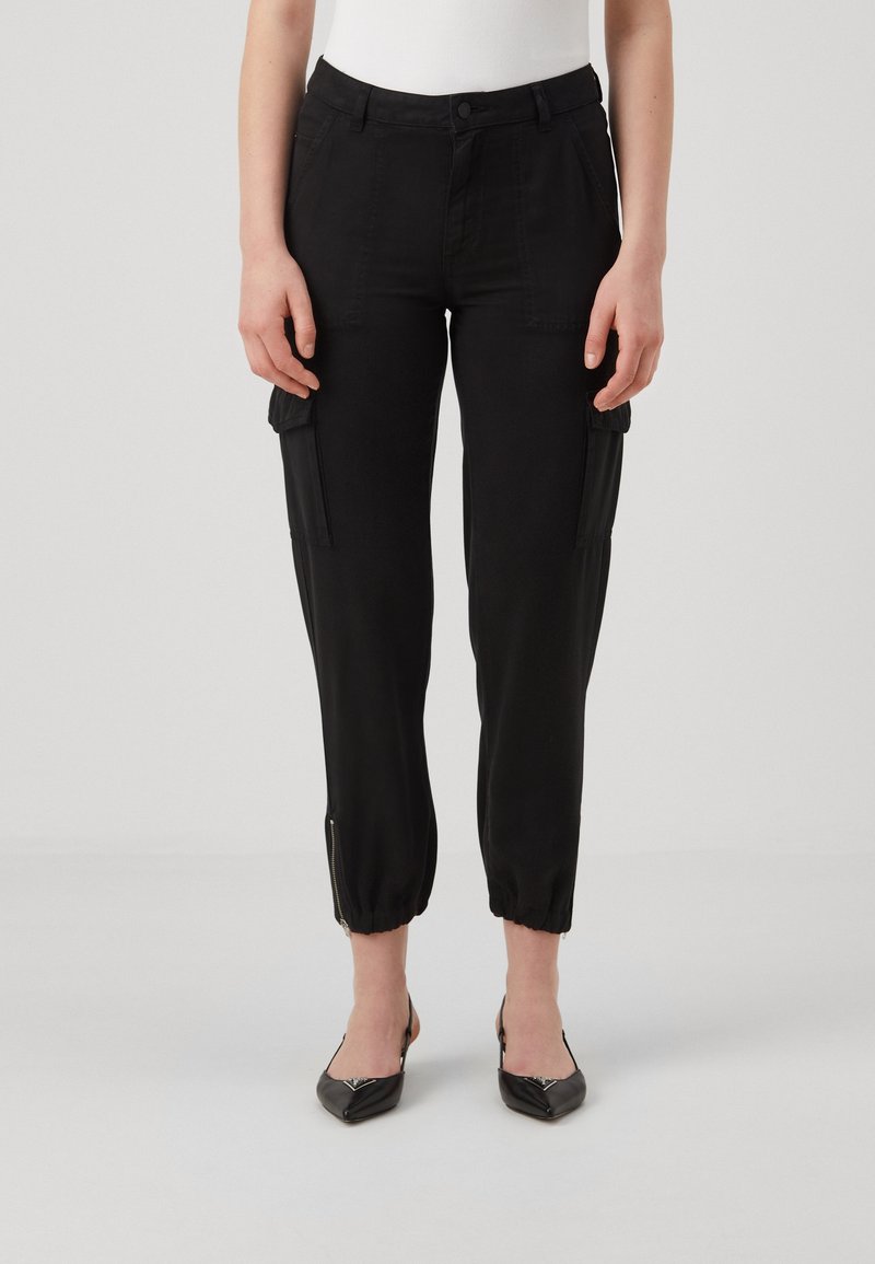 Femme portant un pantalon cargo noir raccourci avec des poignets élastiques et des talons slingback noirs à pointe pointue, debout devant un fond uni.