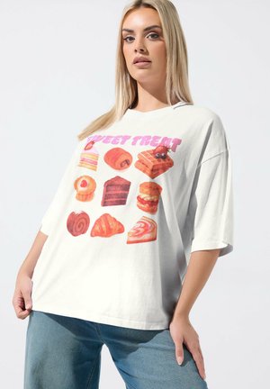 Lyshåret kvinde iført oversized hvid T-shirt med farverige illustrationer af bagværk og teksten "SWEET TREAT" i pink, kombineret med blå jeans.