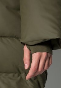 Manica di giacca imbottita verde oliva con polsino a costine. Mano che afferra il tessuto, mostrando una texture liscia e linee pulite.