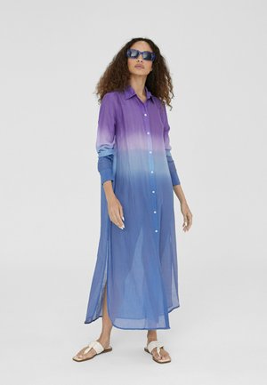 Vestido largo tipo camisa en tejido degradado de púrpura a azul, con botones en la parte delantera, mangas largas y aberturas laterales. Llevado con sandalias blancas y gafas de sol.