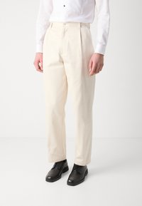Pantalones color crema con una textura suave, cintura alta, frente con pliegues y corte recto, combinados con zapatos negros de cordones.