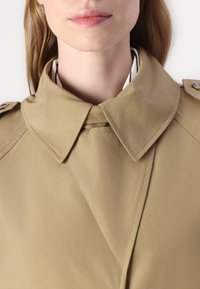 Beige trench coat med stor spredt krage, synlig søm og en glatt, jevn tekstur. Har knappedetaljer på skuldrene.