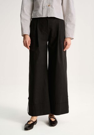 Osoba nosząca czarne spodnie z wysokim stanem i szerokimi nogawkami, zestawione z czarnymi balerinami i jasnoszarym, teksturowanym żakietem.