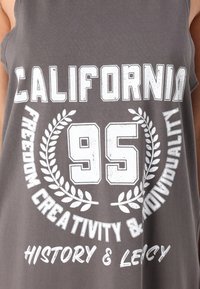Débardeur gris avec des lettres blanches audacieuses. Le texte comprend "CALIFORNIA", "95" et un motif décoratif de laurier, fabriqué en tissu doux.