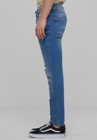 Közepesen kék farmer skinny farmernadrág, elhasználódott részletekkel. Szabványos zsebekkel és testre simuló siluettel rendelkezik, fekete sportcipővel párosítva.