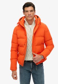 Pomarańczowa, pikowana kurtka z kapturem, z zamkiem błyskawicznym i napami. Noszona na jasnym swetrze w warkocz i niebieskich dżinsach.
