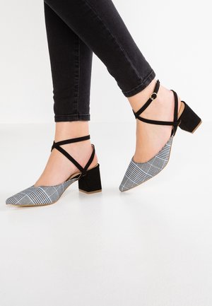 Pieds portant des talons blocs à bouts pointus à carreaux noir et blanc avec des brides croisées noires à la cheville, associés à un jean skinny noir.