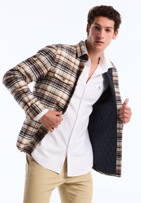 Geruite jas in bruin, crème en marineblauw met een ontwerp van dubbele zakken. Open gedragen over een wit overhemd met knopen. Gequilte marineblauwe voering zichtbaar.