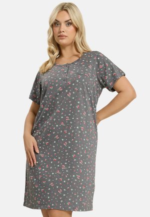 Vrouw draagt een grijze, knielange, korte mouw jurk met een klein bloemenpatroon en een voorzak op de borst, poseert met één hand op de heup.