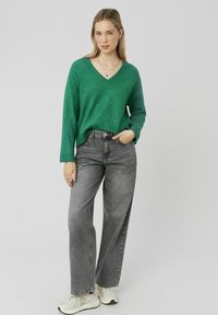 Pull-over vert à col en V en matière texturée, associé à un jean gris taille haute et des baskets blanches, se tenant devant un fond neutre.