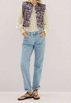 Frau trägt eine cremefarbene Bluse mit Blumenmuster, eine gemusterte dunkle Weste, hellblaue Jeans und schwarze, mit Nieten verzierte Sandalen, die Hände in den Taschen.