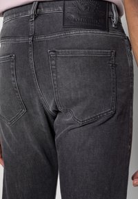 Los jeans de mezclilla negra presentan un acabado desgastado, cinco bolsillos y una etiqueta de cuero con la marca en la parte trasera de la cintura. La tela tiene una textura suave.