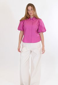 Moteris su ilgais blondinų plaukais, vilkinti ryškiai rožinę palaidinę su pūstomis rankovėmis ir platėjusias baltas kelnės, stovinti priešais paprastą baltą foną.