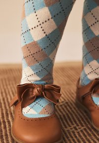 Chaussures en cuir marron à bout rond avec un nœud noué, associées à des collants à motif losanges bleus, bruns et beiges, reposant sur une surface texturée.