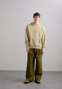 Beige kapucnis pulóver kenguruzsebbel, lazán illeszkedő olívazöld nadrággal és fekete lábbelivel párosítva. Sima textúrák, laza szabás.
