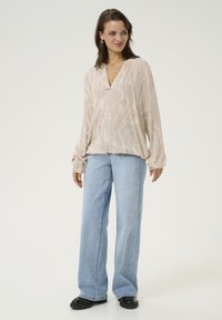 Blouse beige à manches longues avec un motif ondulé, associé à un jean large bleu clair et des chaussures noires. La blouse a un décolleté en V et des manches froncées.