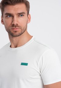 Bílý krátký rukáv tričko s kulatým výstřihem, kontrastním zeleným prošíváním a zelenou obdélníkovou nášivkou na levém hrudníku.