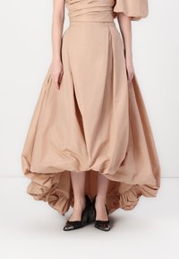 Gaun beige muda dengan bodice yang terstruktur, rok asimetris yang mengalir, dan kain berkumpul di bagian hem. Dipadukan dengan sepatu hak tinggi hitam.
