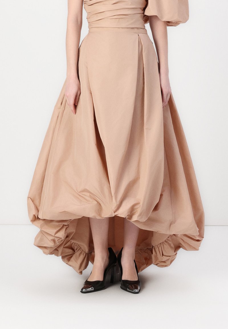 Gaun beige muda dengan bodice yang terstruktur, rok asimetris yang mengalir, dan kain berkumpul di bagian hem. Dipadukan dengan sepatu hak tinggi hitam.