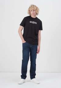 Juodas medvilnės marškinėliai su balta "GUESS" logotipu, derinami su tamsiai mėlynomis džinsais ir baltais sportbačiais. Modelis pozuoja atsipalaidavusia poza.