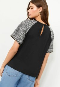 Sort t-shirt med teksturerede, mønstrede grå ærmer. Har en nøglehulåbning bagpå og en glat overflade. Sammen med blå jeans.