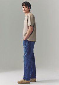 Hellbeiges Baumwoll-T-Shirt, locker geschnitten, kombiniert mit blauen Denim-Jeans. Die Schuhe sind hellbraune Wildleder-Slipper. Schlichtes Design, ohne Muster.