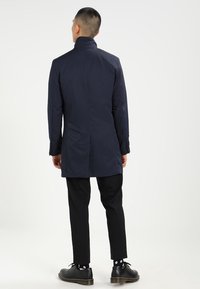 Cappotto lungo navy con colletto alto, polsini con bottoni e spacco laterale. Indossato sopra pantaloni neri e scarpe con suola texturizzata, con pois bianchi.