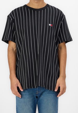 Mann trägt ein schwarzes kurzärmeliges Hemd mit weißen vertikalen Streifen und kleinem Tommy Jeans-Logo auf der Brust, kombiniert mit blauen Jeans.