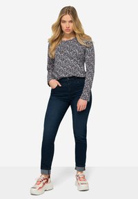 Top de manga larga con un patrón en azul marino, combinado con jeans oscuros. La modelo lleva zapatillas blancas y naranjas de suela gruesa. Jeans de pierna recta con dobladillo.