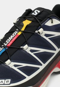 Μαύρο και ναυτικό παπούτσι τρεξίματος με υφασμένο επάνω μέρος, λευκά γεωμετρικά σχέδια, κίτρινες και κόκκινες λεπτομέρειες και ένα ασφαλές σύστημα κορδονιών.