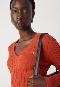 V-neck, rust-colored knitted sweater with cable pattern and small logo. Model holds a black shoulder bag with a smooth texture.