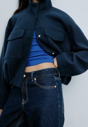 Personne portant une veste courte bleu marine foncé sur un crop top bleu avec un jean en denim foncé, main glissée dans la poche du jean.