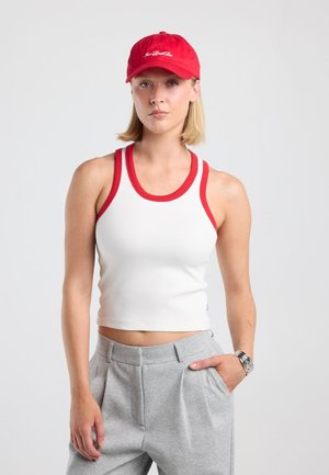 Femme debout, portant une casquette rouge, un débardeur blanc à liseré rouge et un pantalon gris, une main dans la poche devant un fond uni.