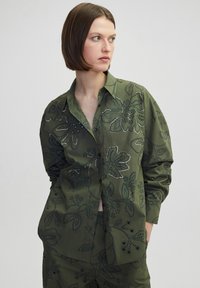 Olijfgroene button-up shirt met bloempatroon borduurwerk en zilveren draadaccenten. Beschikt over een klassieke kraag en losse mouwen.